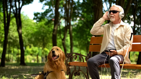 A visually impaired man wearing headphones is sitting on a park bench with his dog beside him.